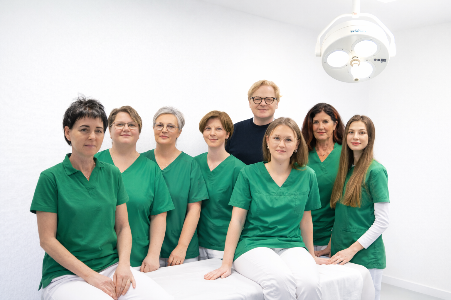 Das vollstaendige Praxisteam der Orthopaedie Dr. Bastian: Drei Aerzte in weissen Kitteln und 7 Praxismitarbeiterinnen stehen gemeinsam vor dem Praxiseingang und laecheln freundlich in die Kamera