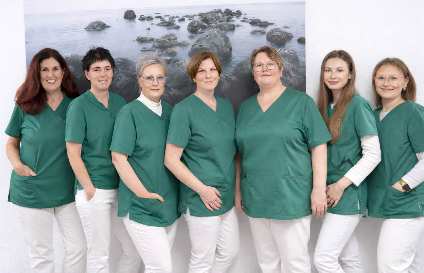 Das vollst&auml;ndige Praxisteam der Orthop&auml;die Dr. Bastian steht und sitzt gemeinsam vor dem Praxiseingang: sieben Praxismitarbeiterinnen